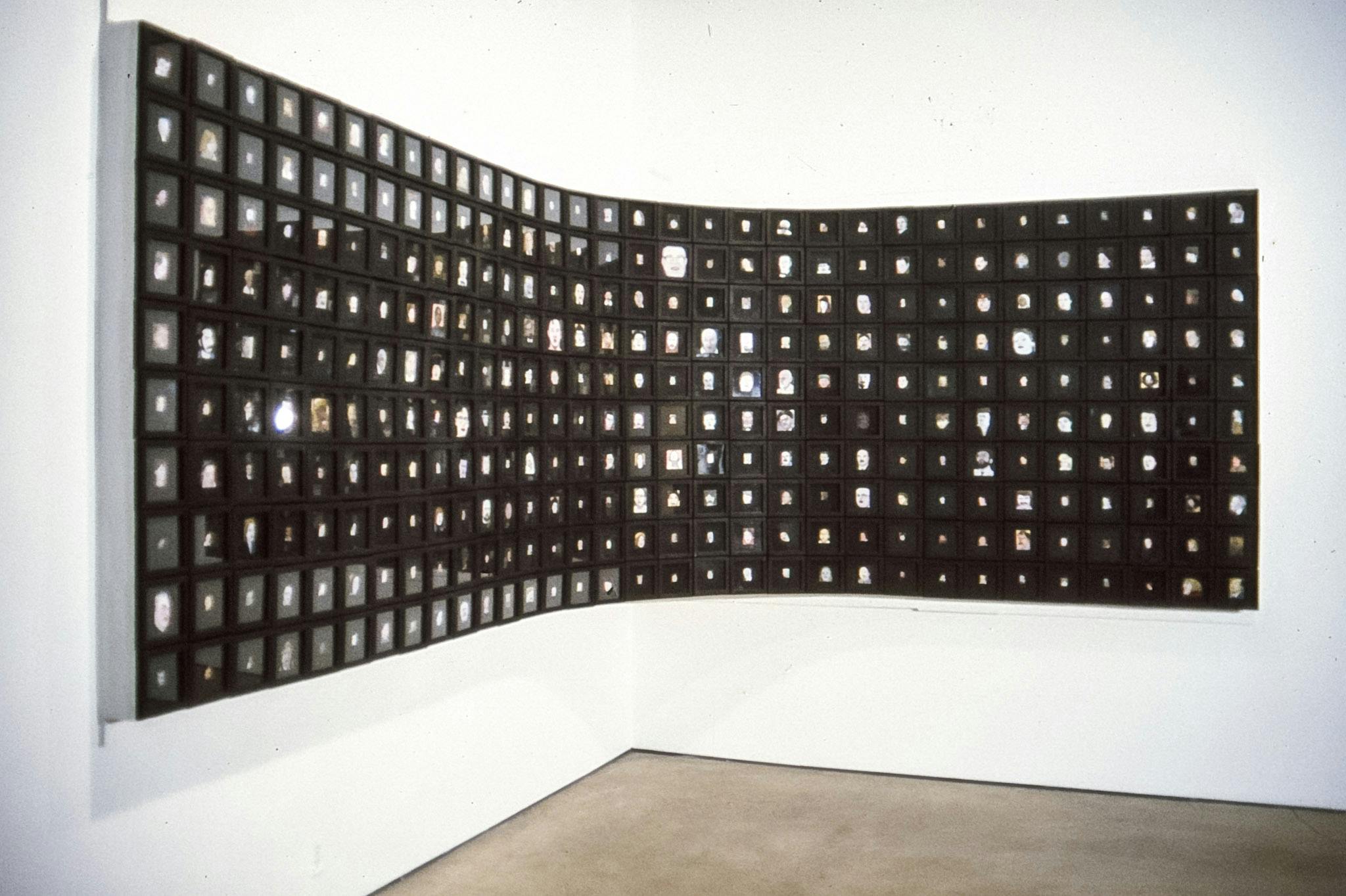A photo collage of 320 small photographs of human faces mounted at a corner of the gallery space. Each photograph is black framed; some images are colored and some are black and white. 