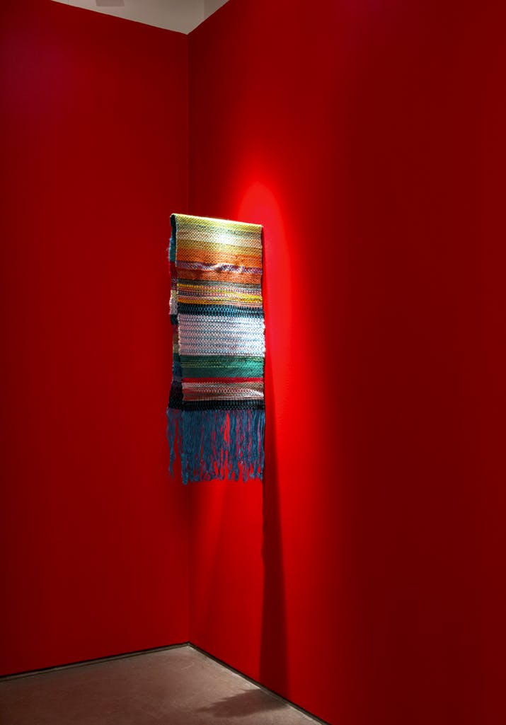 A scarf-like textile hangs from a bar attached to a gallery wall. The textile has a colourful stripe-pattern and blue tassels. The gallery walls are painted in red, and the floor is light grey. 
