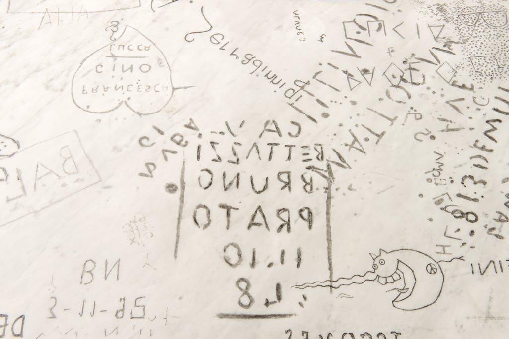 Detail image of the surface of a white marble bench sculpture. Various carved graffiti marks, Roman alphabet characters, Arabic numbers, a heart, squares, and other marks, cover the surface.