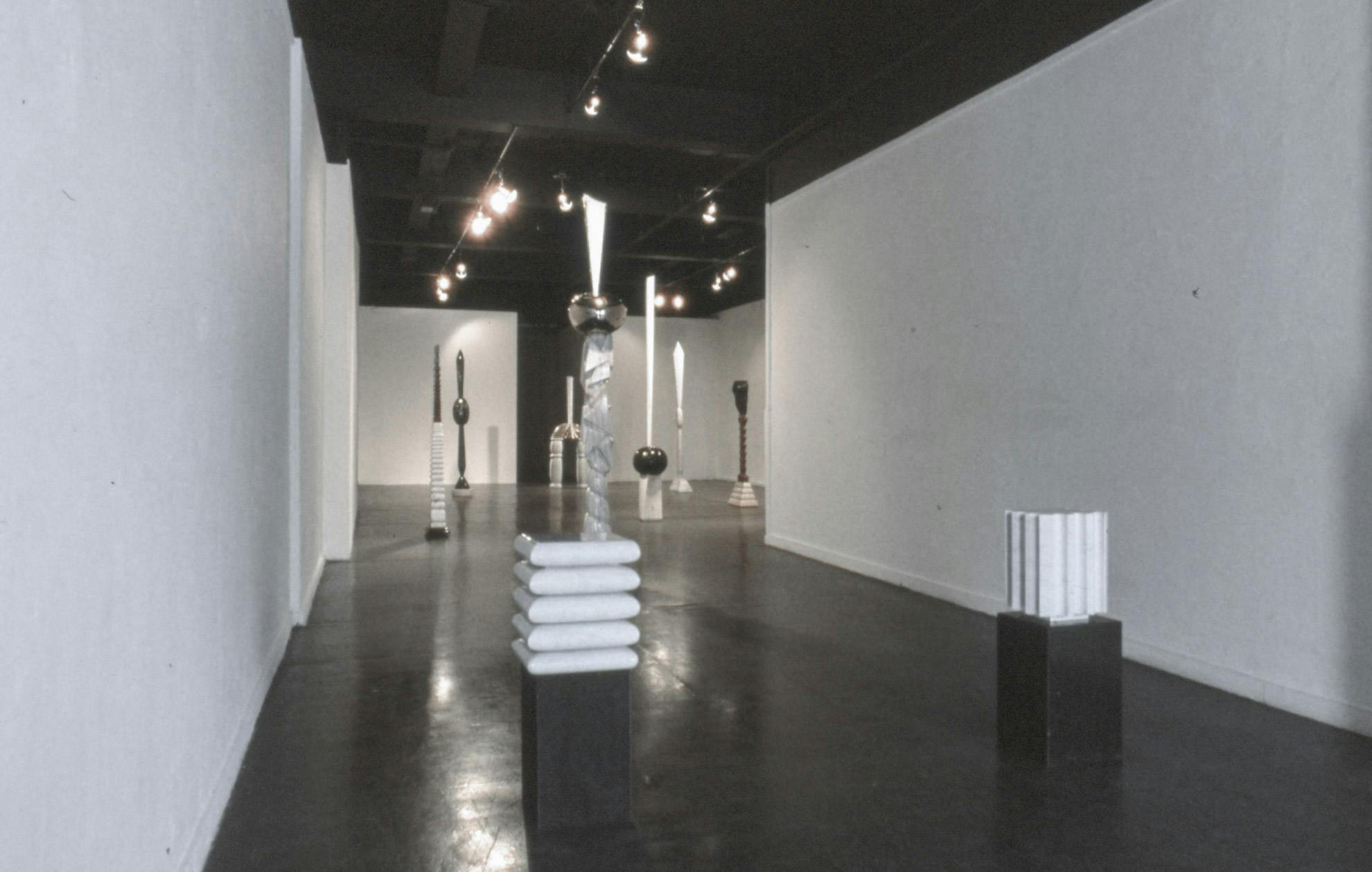 Several sculptures in a gallery. In the foreground, two small white curved forms sit atop black rectangular shapes. In the background the forms are tall and thin, with both bulbous and narrow parts.