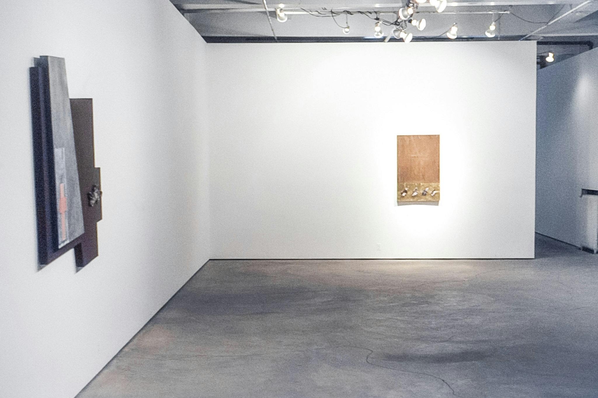 The corner of a gallery space. On one wall, there is a wood panel with gold foil and small bits of material along its bottom edge. On the other, an asymmetrical work with layered painted panels.