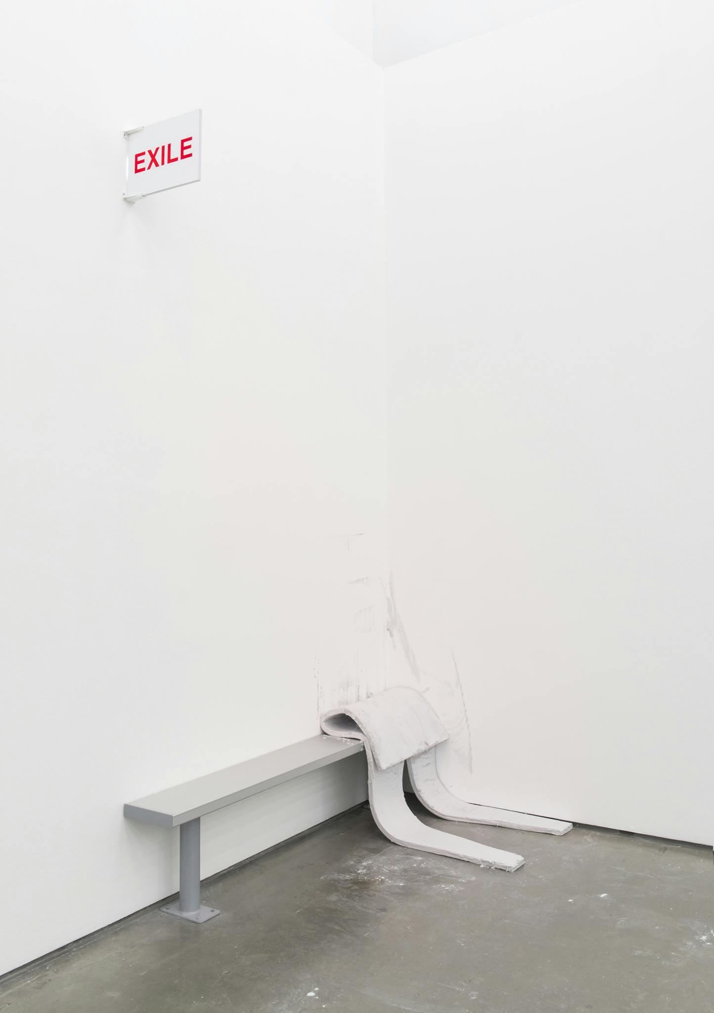 An aluminum bench is installed in the corner of a gallery. On it sits a thin, slumped foam cutout in a human-like form. An exit sign reading EXILE instead of exit hangs on the wall above.