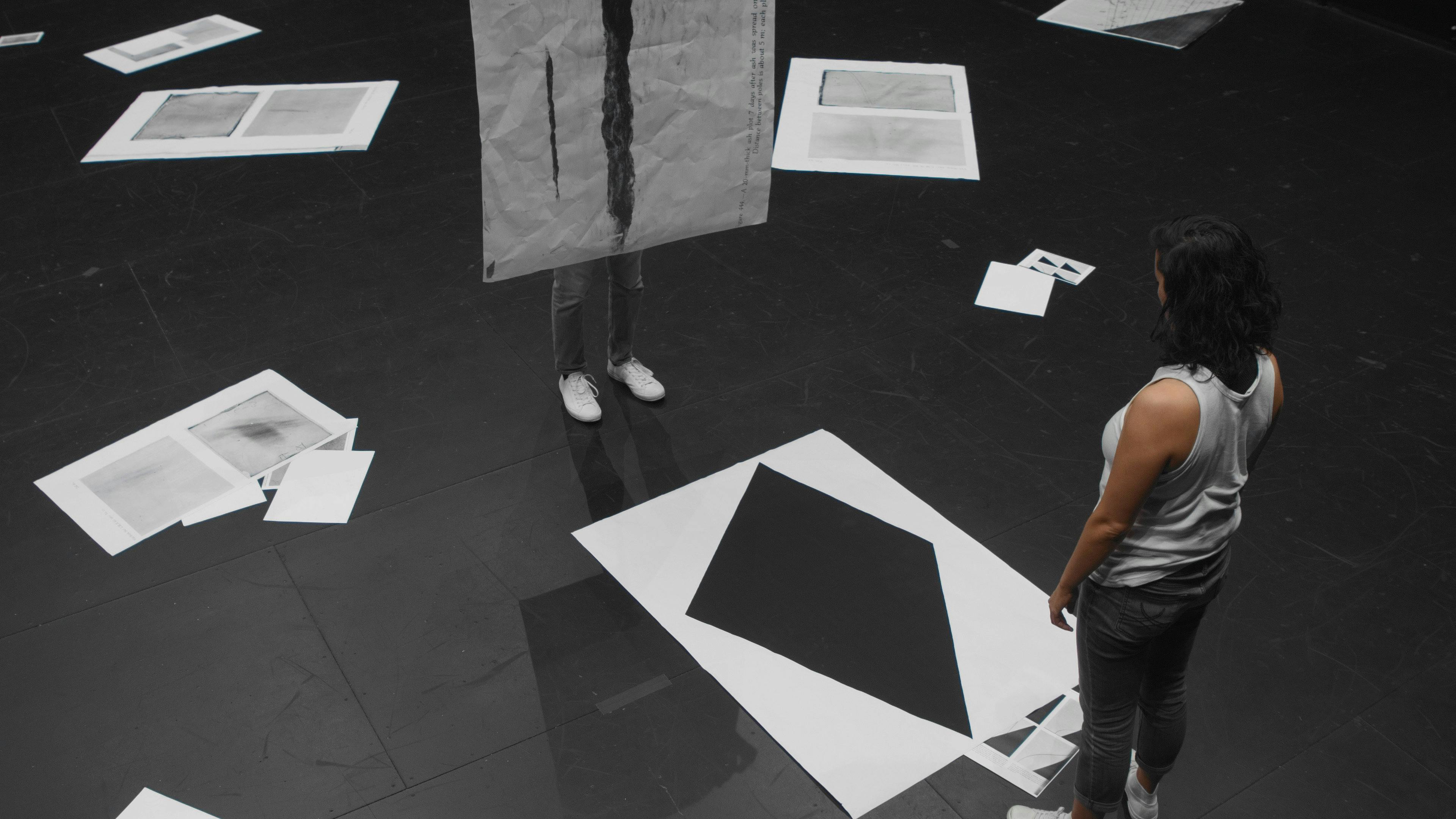 An aerial view of a person with a black hair standing in front of an artwork placed on the floor. The work is a large paper with a diamond- shaped cut-out in the middle.