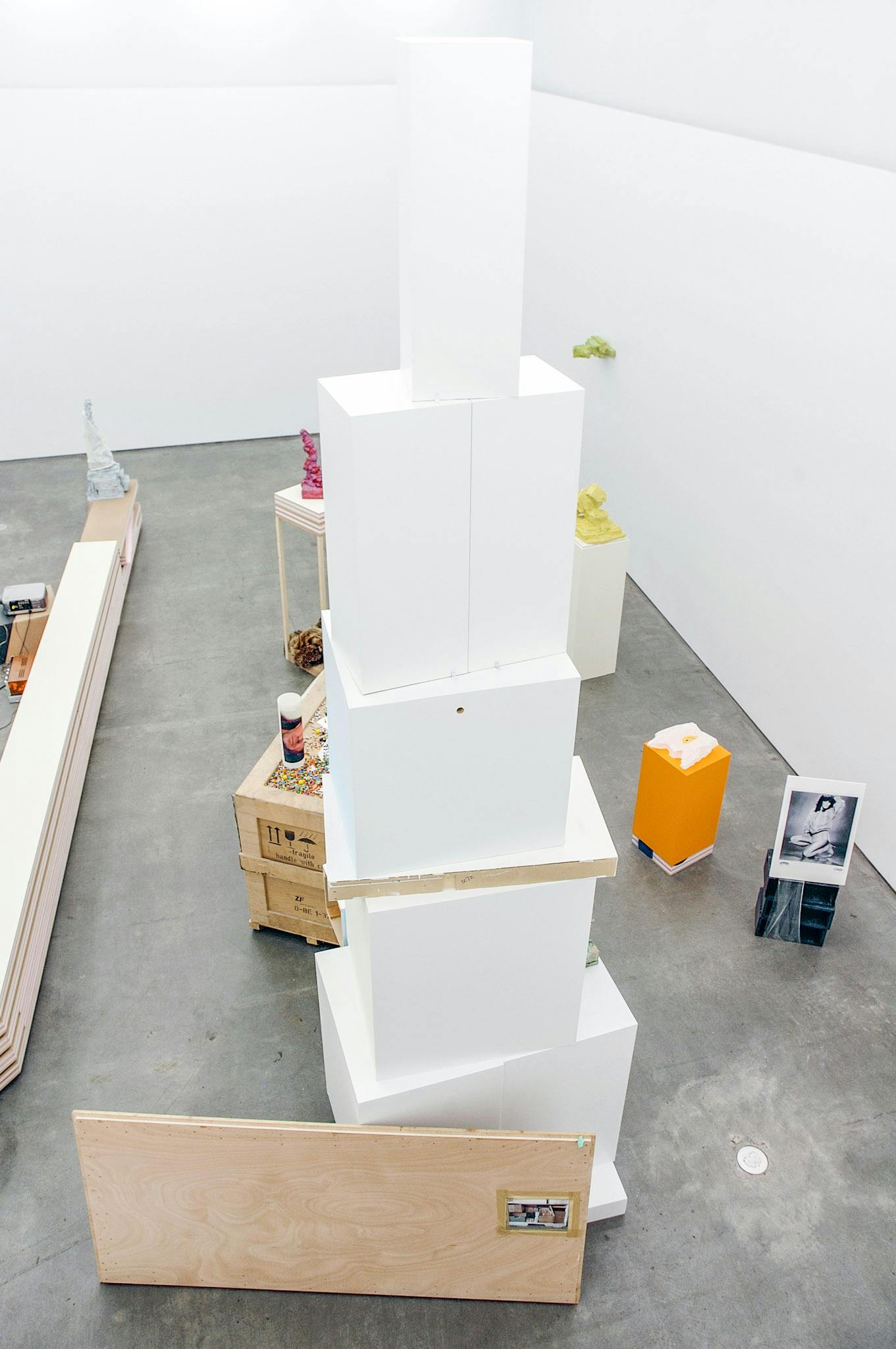 A view from above of artworks in a gallery. At the centre, there is a tall tower of several white plinths, stacked. Other objects include planks of plywood and a photo of a person, posing semi-nude.