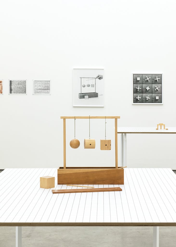 A wood object installed on a white table. Three differently-shaped blocks hang from a simple frame. A wood cube and two other wooden pieces lay in front of the frame. 