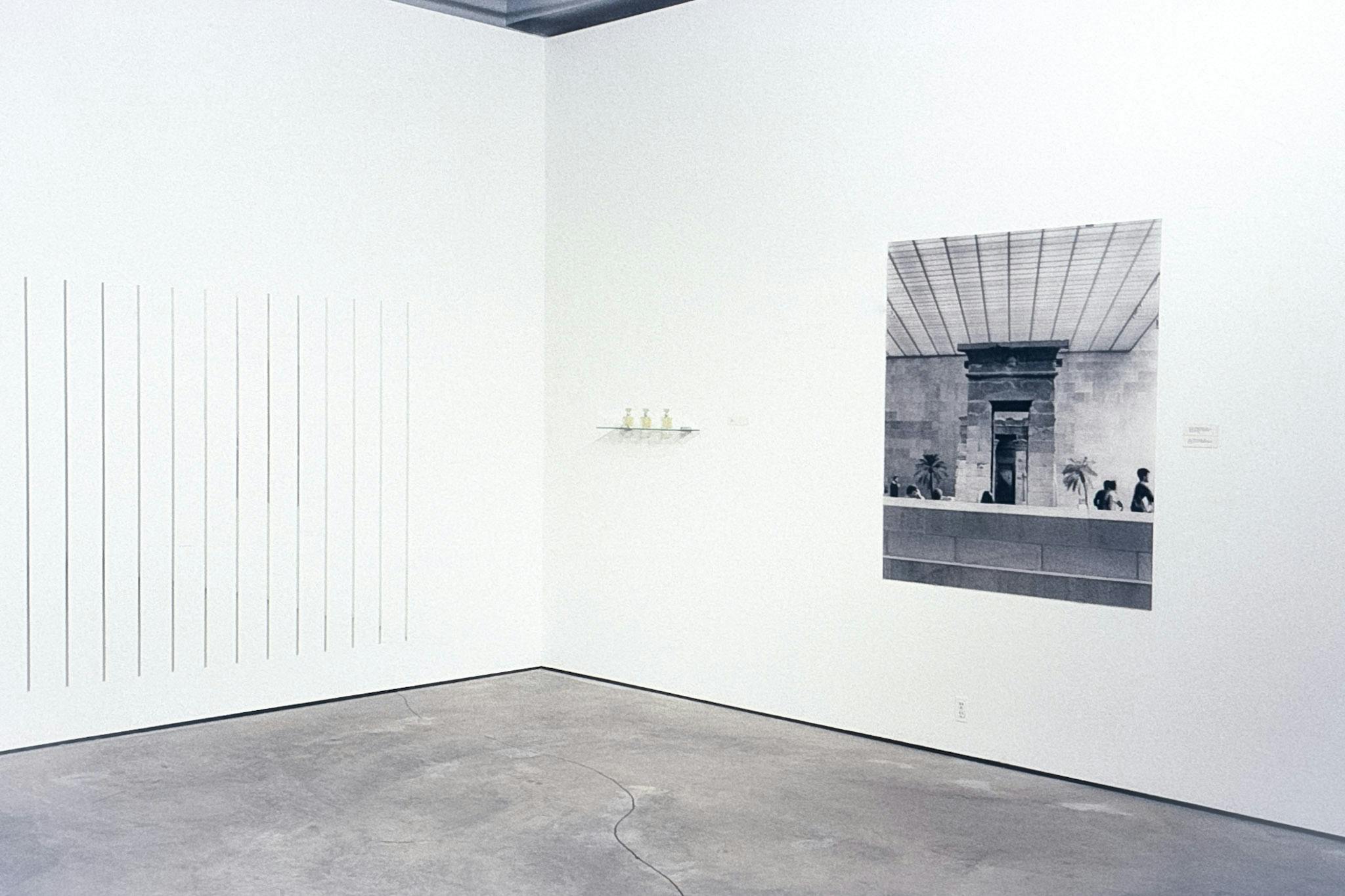 3 artworks in the corner of an art gallery. One is dark slits on a wall, one is small bottles on a glass shelf, and one is a large black and white photo of a room with brick walls and tiled ceilings. 