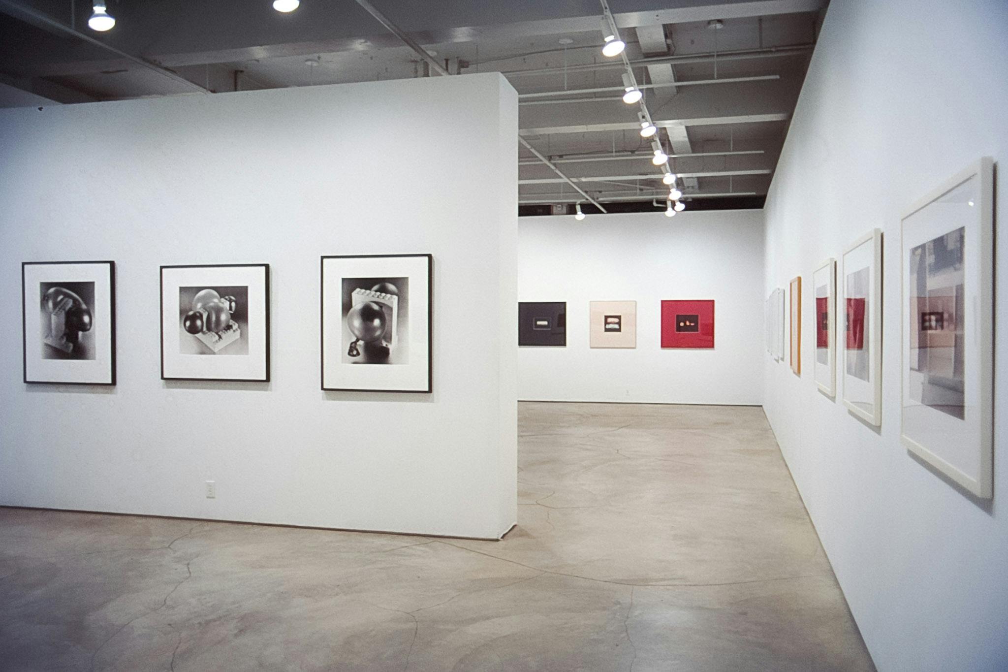 Eleven art pieces mounted on three gallery walls are visible in this image. Three black and white photographs on the front wall depict silver organic shapes being attached to the white blocks. 