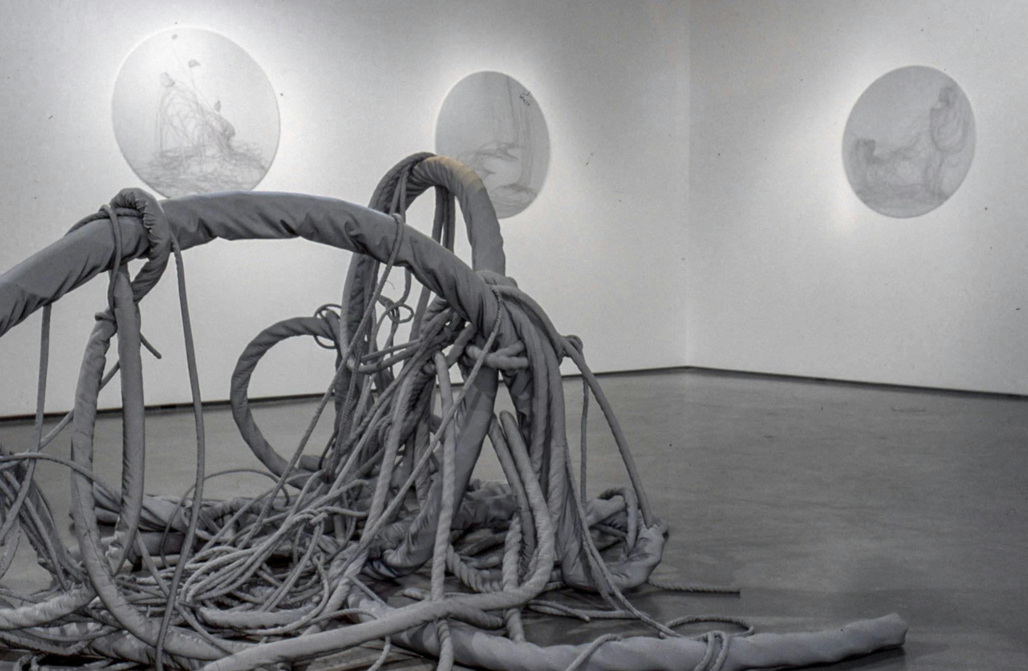 A large grey sculpture that looks like a tangled fishing net is placed in the middle of the gallery floor. Three circular shaped sculptures that resemble white marble are mounted on the walls. 