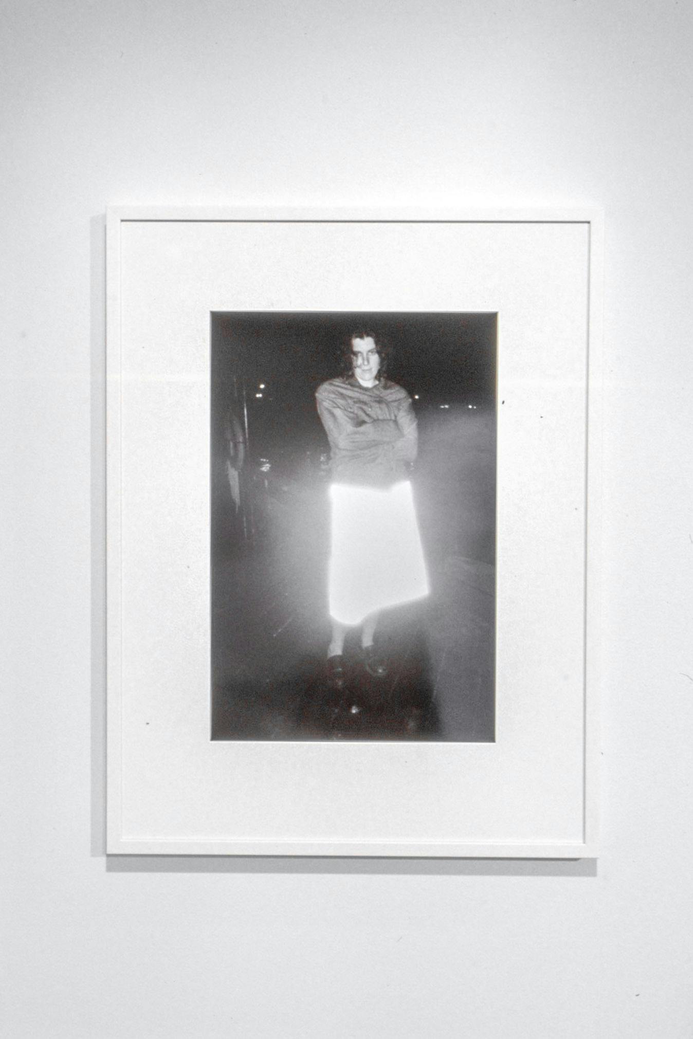 A framed black and white photograph is mounted on a wall. It depicts the entire body of a woman standing on a street at night. This person wears a skirt that reflects the flashing light of the camera.
