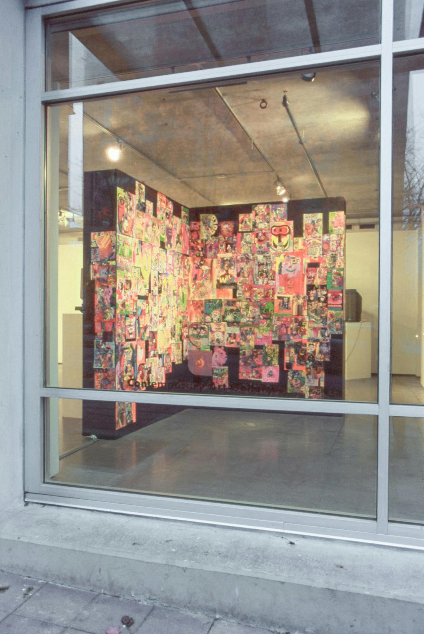 An installation image of artworks in a gallery taken from outside through the windows. A countless number of neon coloured two-dimensional works, mostly drawings and collage pieces, cover two black separation walls.