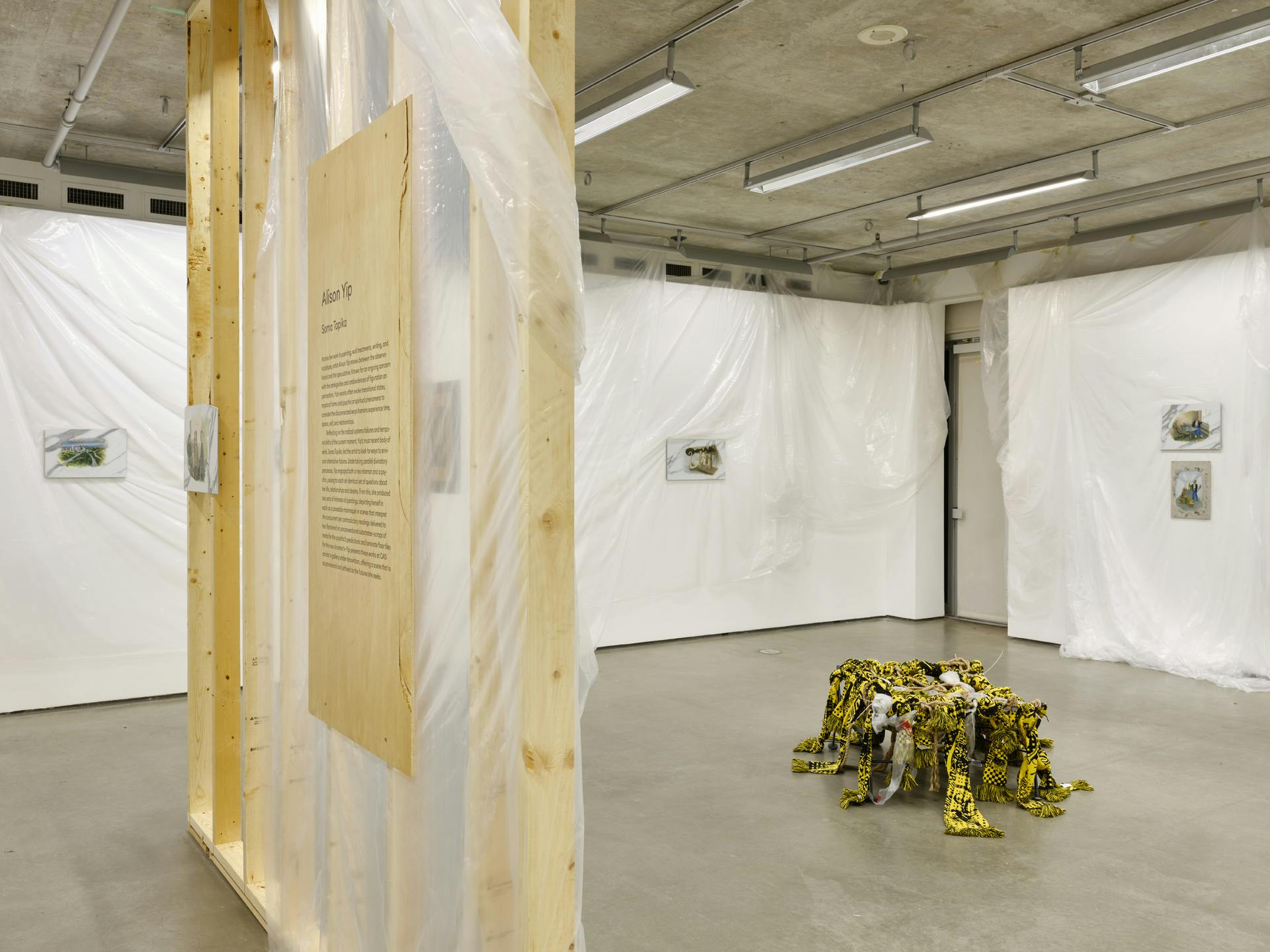  Small paintings hang on gallery walls draped with plastic sheets. An angled view of an unfinished wall structure intersects the view. A sculpture made of textiles, plastic and rope sits on the floor. 