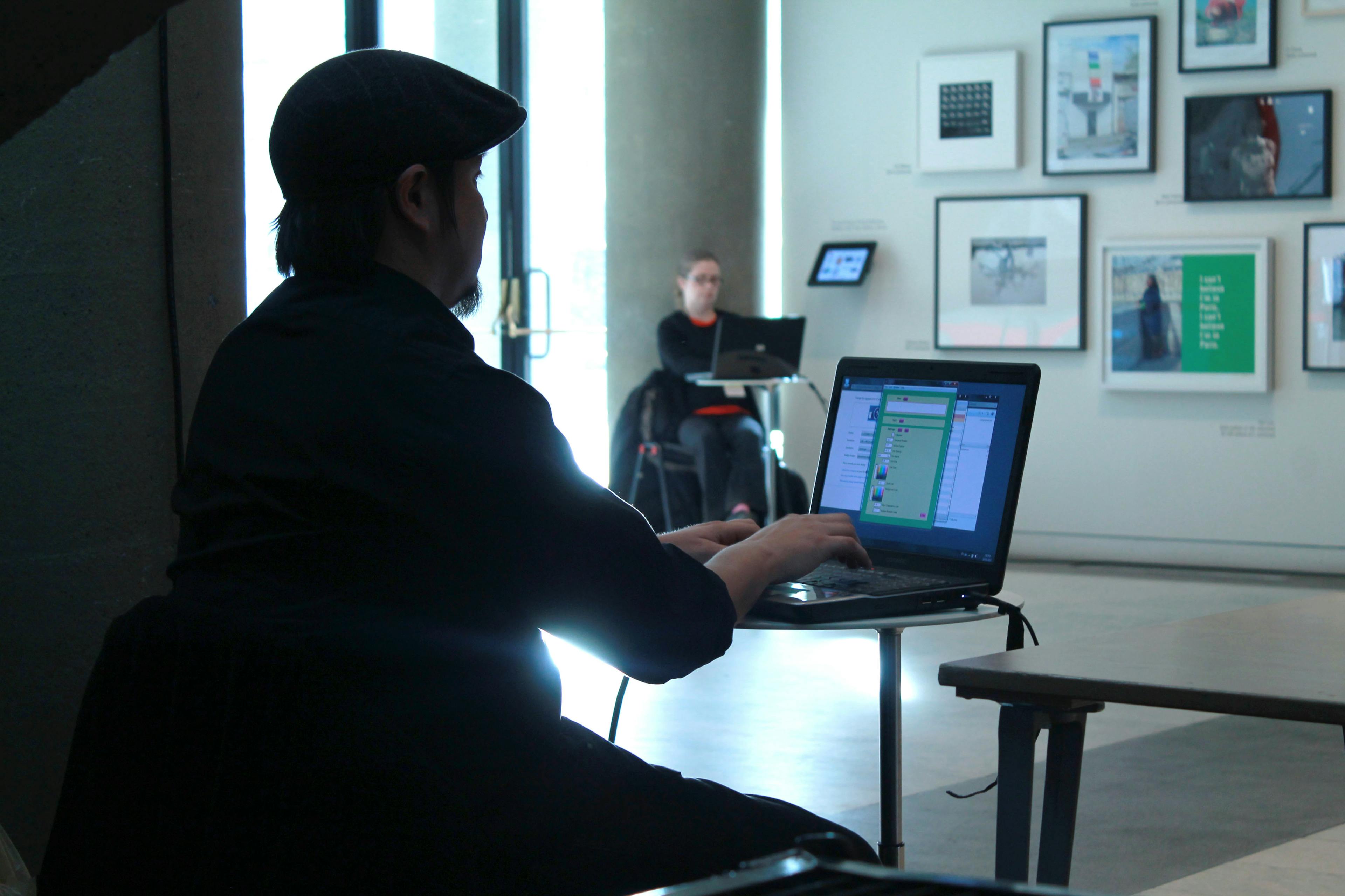 A person sits in front of a laptop at a small circular table. They face a white wall filled with framed artworks. Beside the framed artworks, another person sits with a laptop at a table.