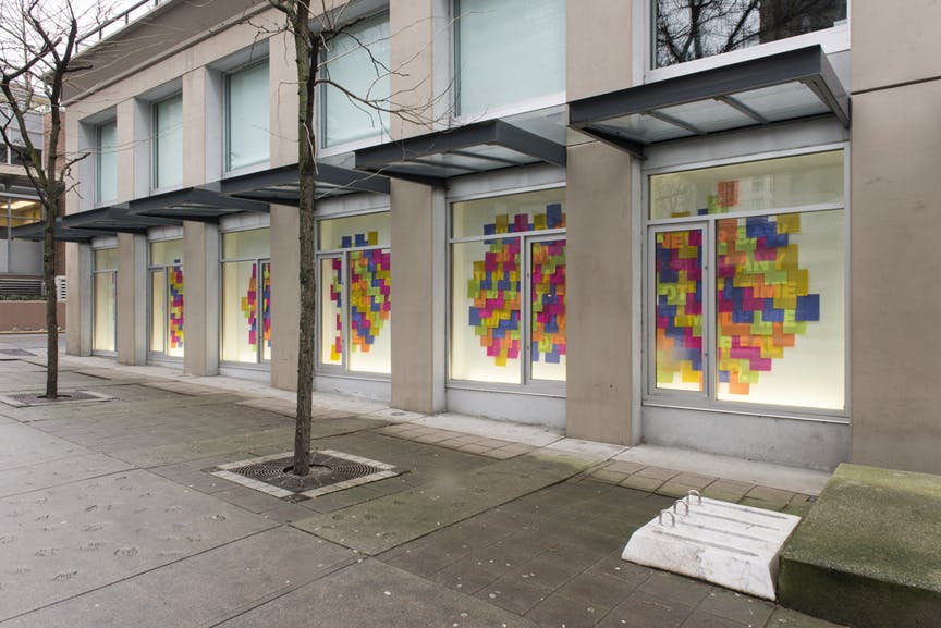 Dozens of coloured sticky notes mounted in windows across CAG’s exterior facade. Semi-transparent Roman letters are visible on these overlapped notes, some of which read "ANY," "NEVER" and "TIME."