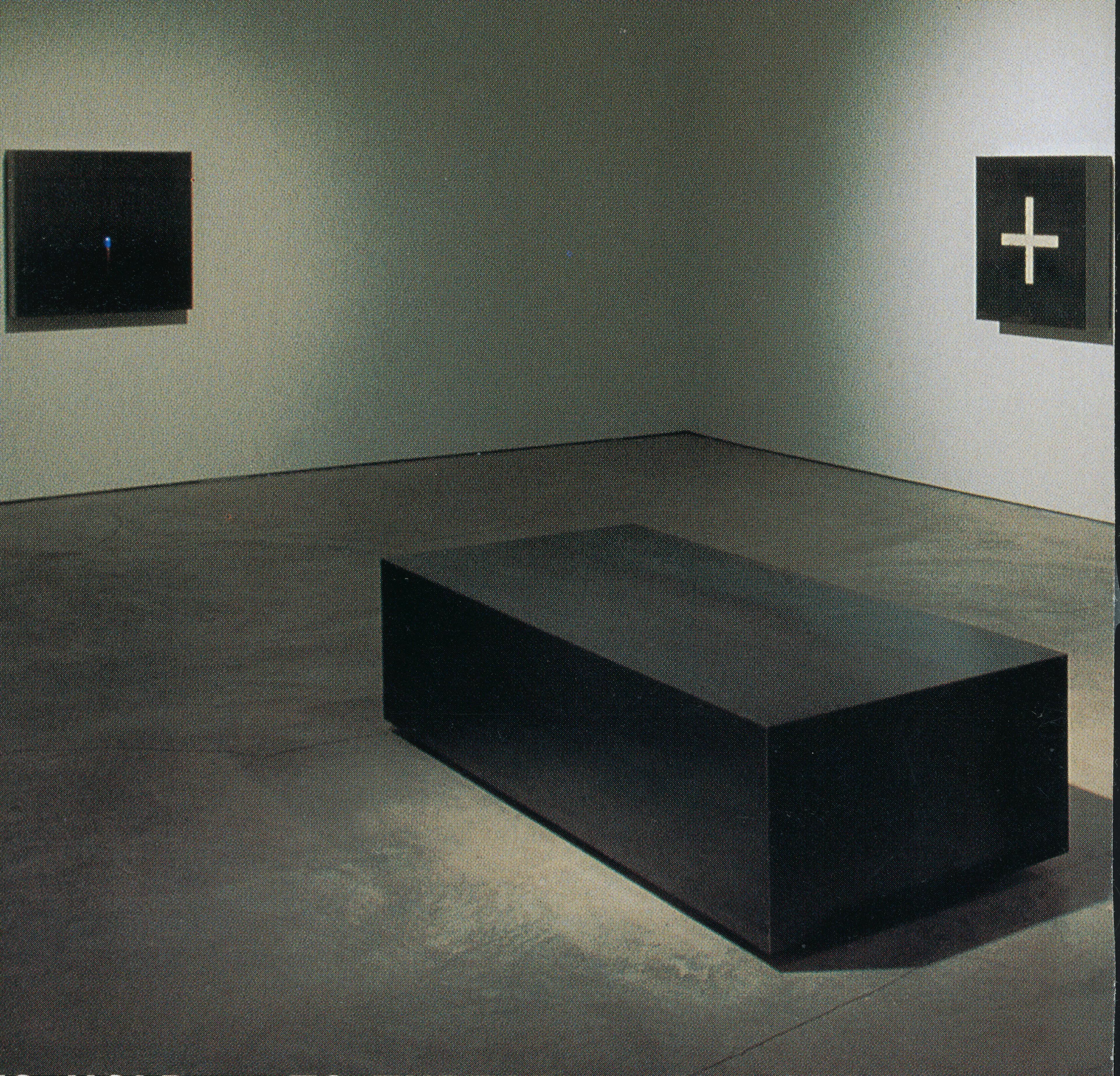 A large steel-made sculpture is placed in the middle of the dimly lit gallery. It is rectangular and has a tarnished dark grey colour. The sculpture is directly placed on the floor. Several other steel-made works are also installed on the walls.