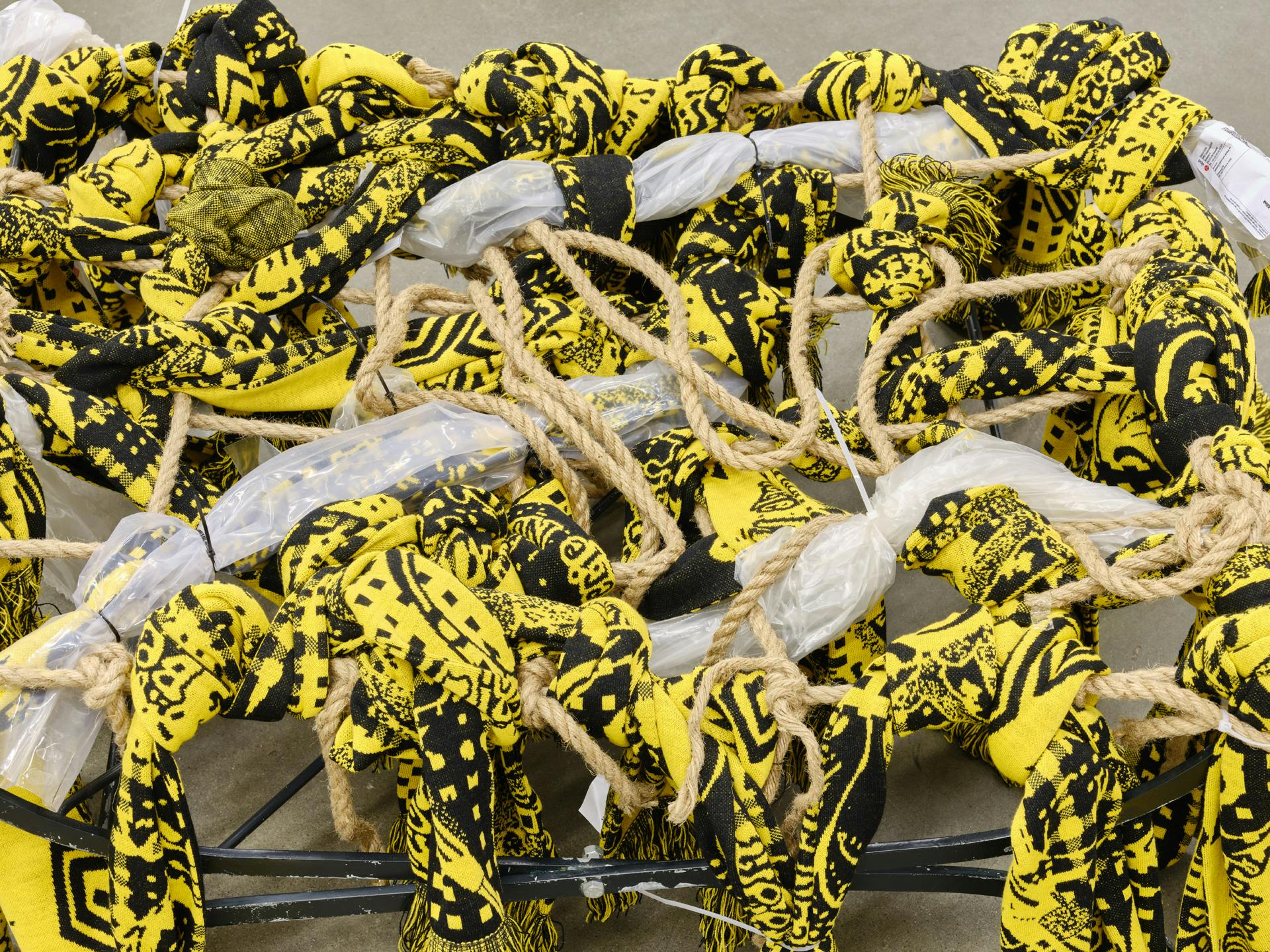 A close view of a sculpture made of yellow and black scarves, brown rope and scraps of plastic packaging tied in knots and draped over a metal seat-like structure. 
