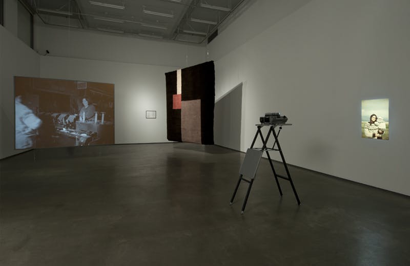 Sarah Browne's artworks installed in a gallery. A film is projected on a large screen next to a large, hand-knotted carpet hanging from the ceiling. A slide projector projects an image on another wall. 