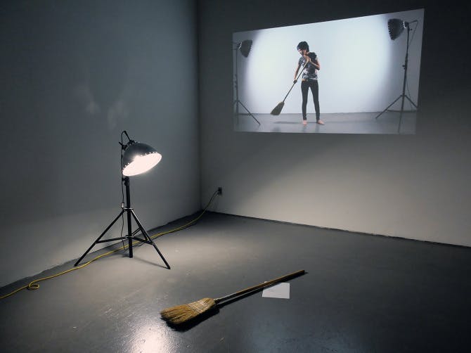 A spotlight and a broom are placed on a gallery floor. A video of a person sweeping with a similar broom is projected onto the white wall. 