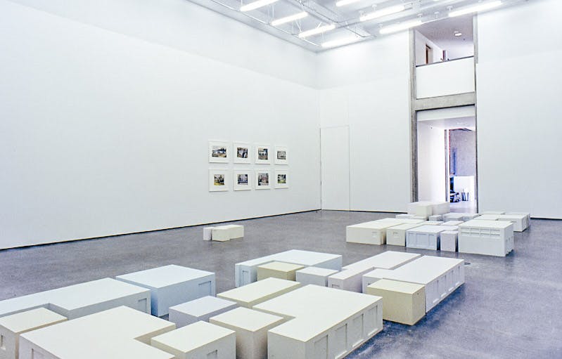 In a gallery space, many pale-coloured cube sculptures are placed on the floor. They are placed to mimic a city landscape. Eight photographs are mounted on the wall near the entrance.