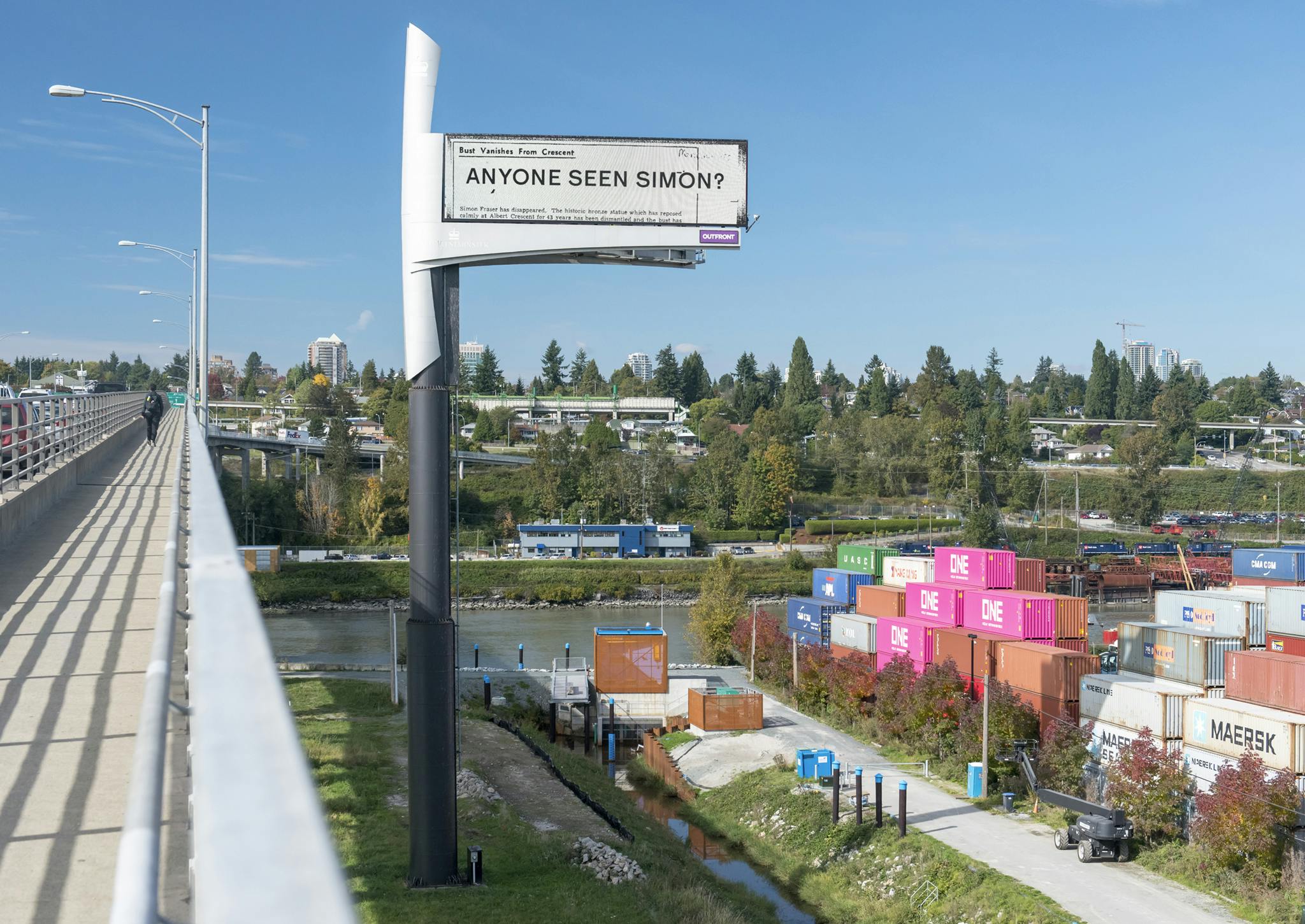 A billboard stands high off the ground with a bridge on one side and shipping containers on the other. The billboard reads “ANYONE SEEN SIMON?” in black text. 