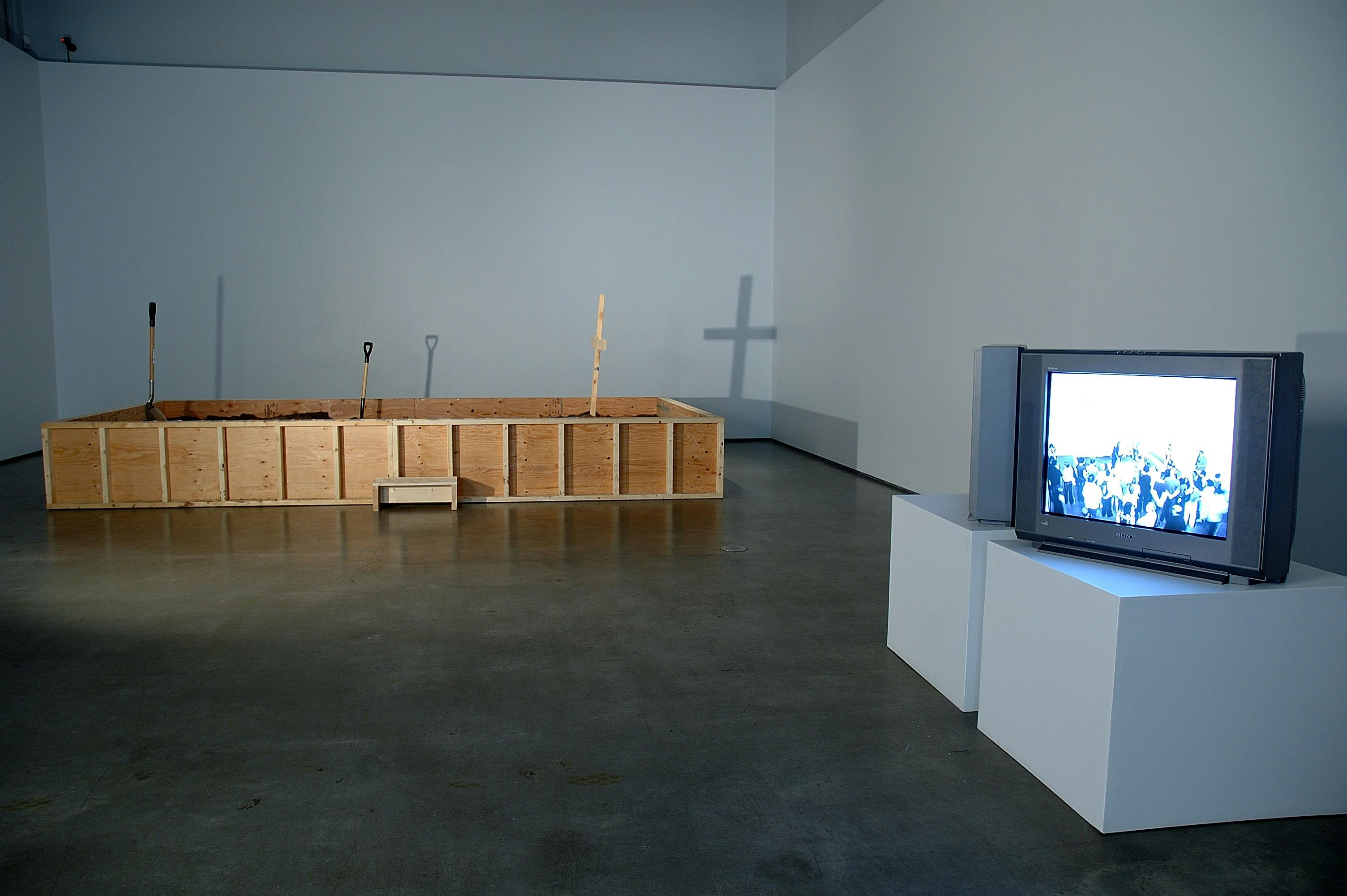 An installation image of several artworks in a gallery. A large unsealed wood box is placed on the floor near the far wall. A CRT TV placed on the pedestal shows the image of an assembly of people.