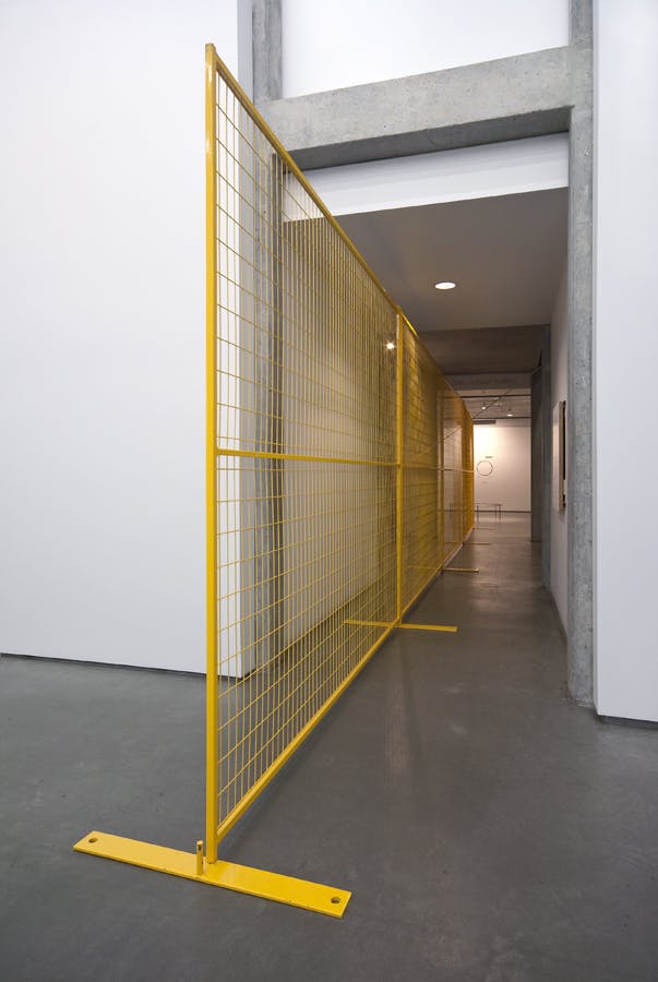 A long, yellow metal fence stands in the middle of a hallway connecting gallery spaces. The fence stands to the side of the hallway, allowing people to pass through.