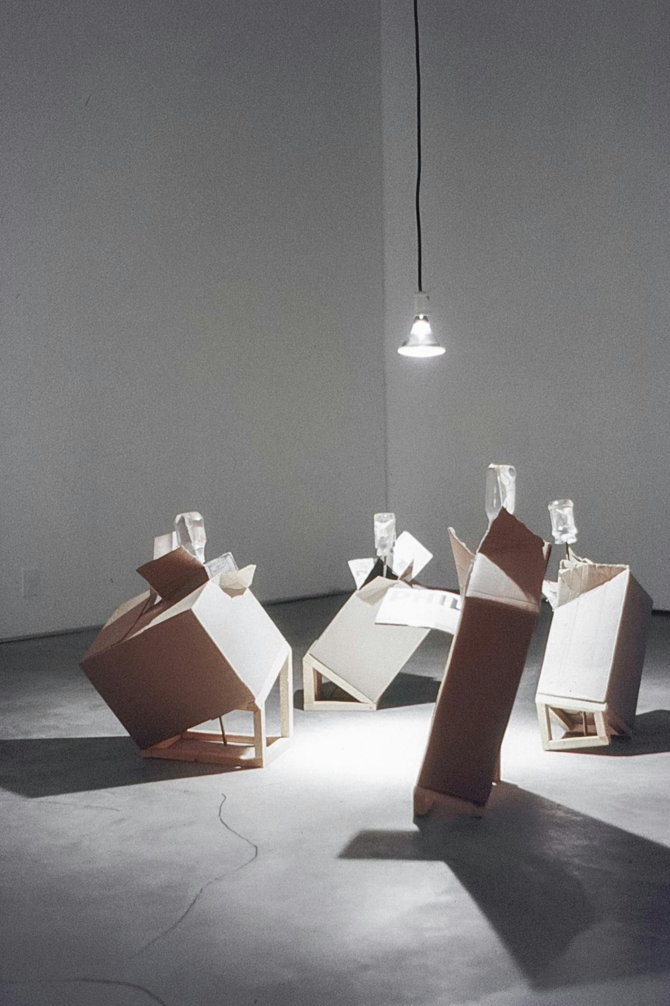 A lightbulb hangs low from the ceiling of a dark gallery space. 4 small sculptures are aglow. They are cardboard boxes, on small wood ramps, with large screwdrivers stuck in their opened corners.