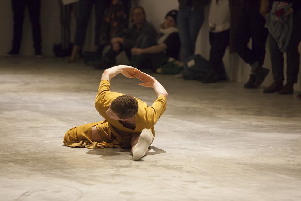 A dancer lays in a partial split on the floor of a gallery space with audience members watching at a distance. Their arms are bent behind them with the tips of their fingers touching.
