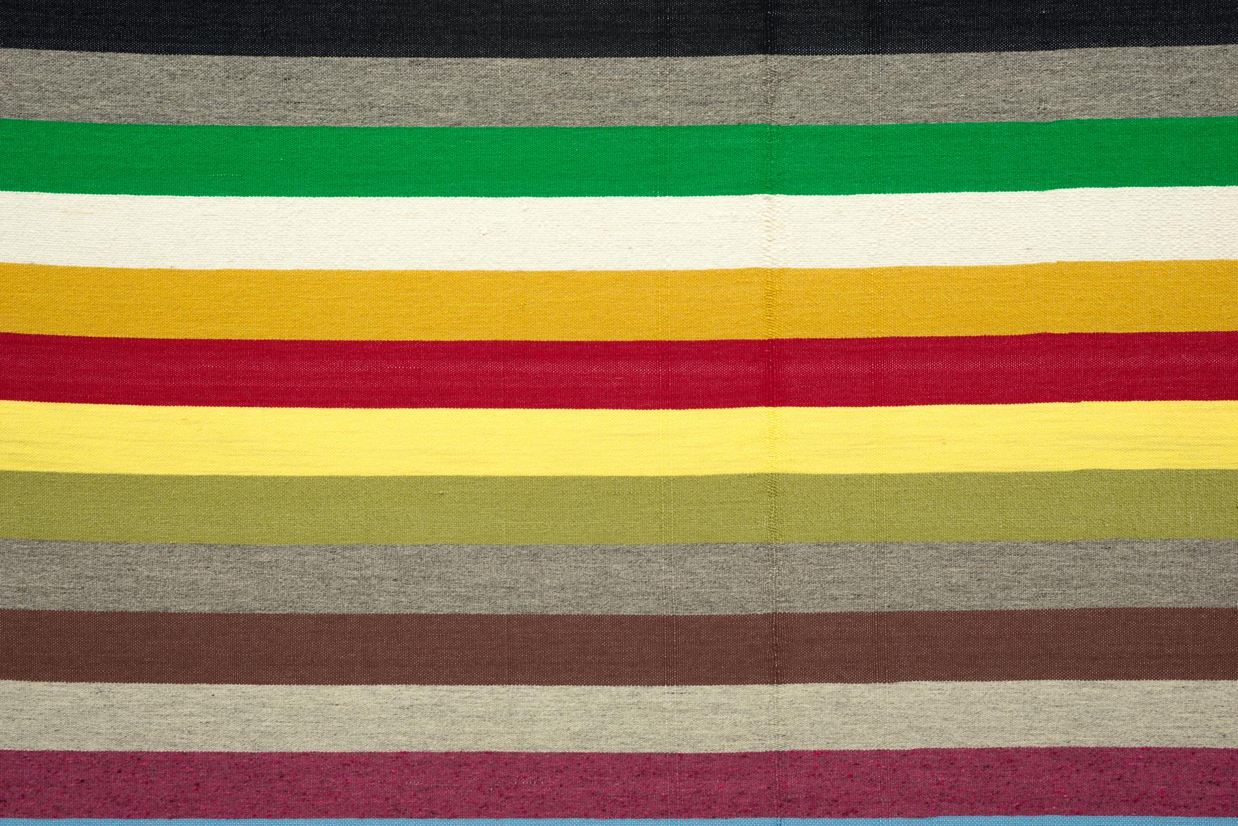 A close up view of a weaving. The weaving is made up of horizontal stripes in various colours including, browns, greens, yellows, greys, white, red and purple.