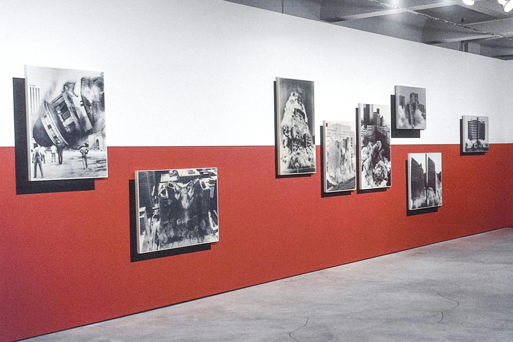 Eight large artworks on a white and red wall. The works are large black and white photos of buildings collapsing. The works are mounted slightly away from the wall, creating dramatic shadows. 