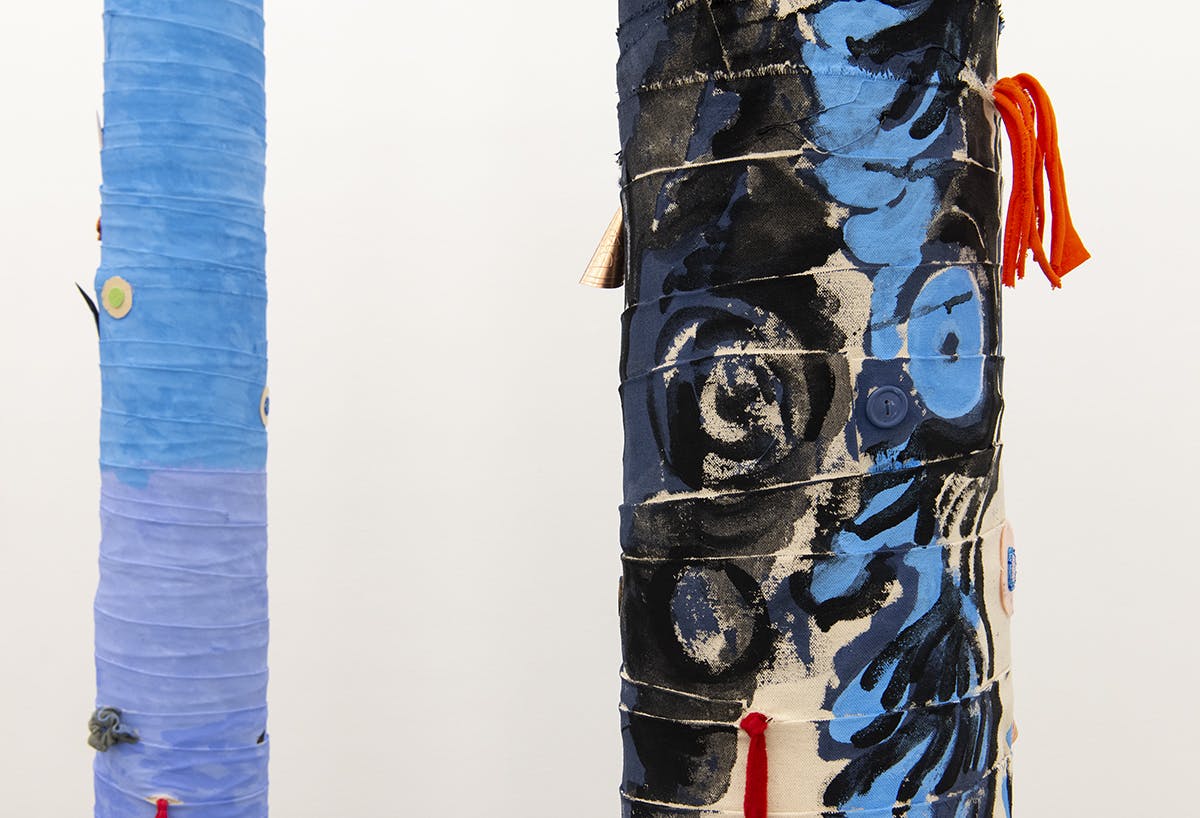 This is a close-up view of Charlene Vickers’ sculptures. Tall cylindrical objects are wrapped in fabric, which is painted blue. Some small objects including buttons are attached to the sculptures.
