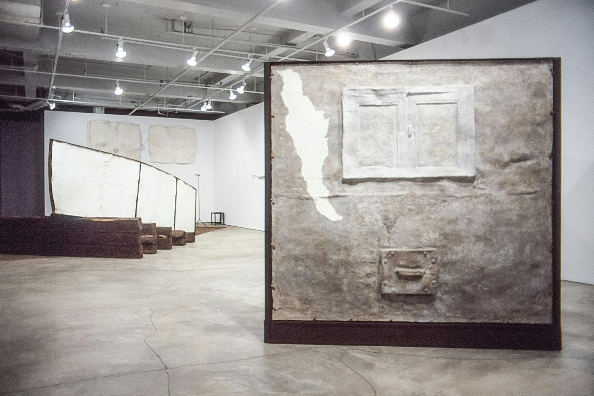 The installation view of the gallery shows three artworks. A pair of white sheets are installed on the back wall, foregrounded by a white boat head. A white cubical-shaped sculpture is in the front.  