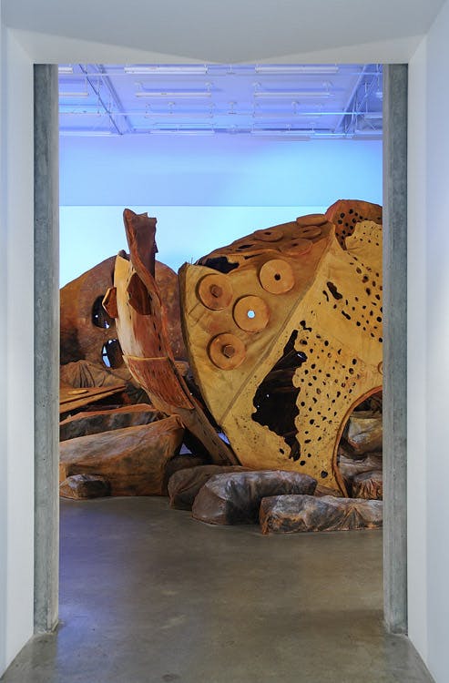 An installation image of a large artwork by Donald Lawrence inside a gallery. An assemblage of distorted and decayed metal parts of some large industrial machine is placed on the floor.