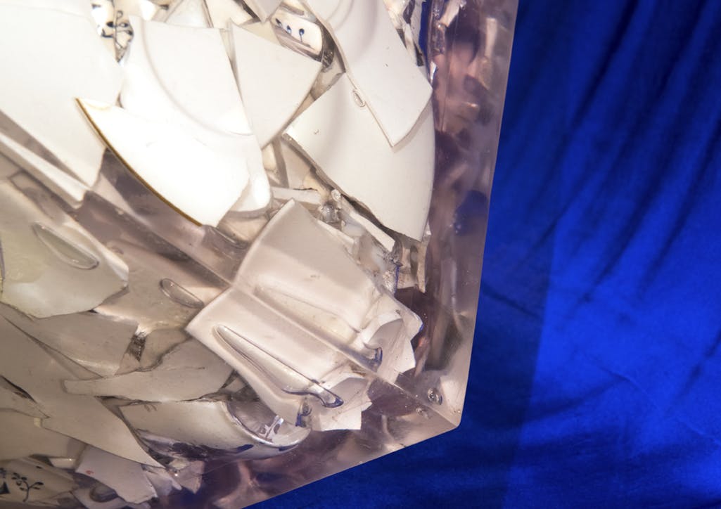 A close view of a clear, block-like sculpture of encased broken ceramic pieces installed on a plinth. A royal blue cloth drapes behind the sculpture. 