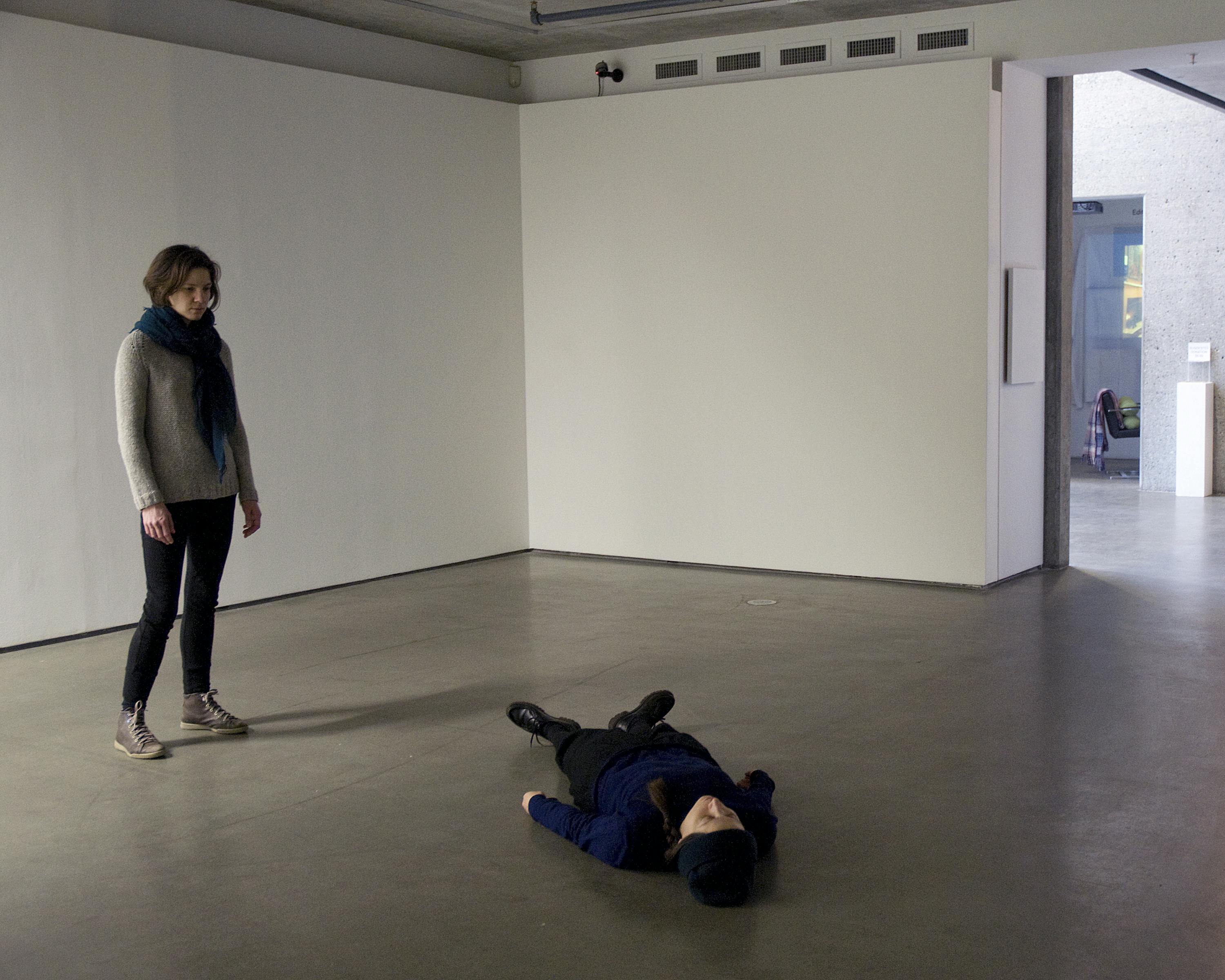 Two people are performing in a gallery space. The one wearing a blue-green scarf is standing straight and looking down at the other, who is lying down on the floor.