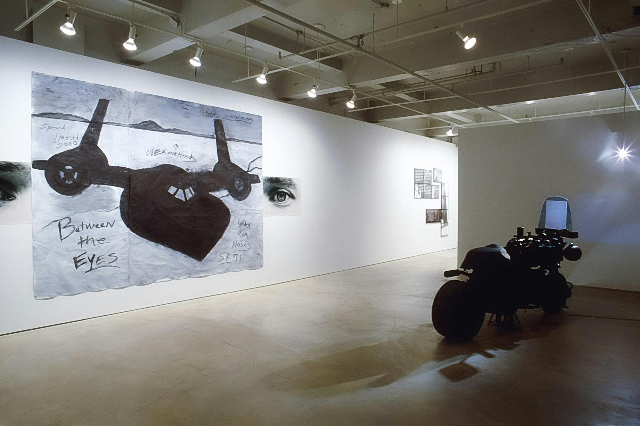 A large drawing is installed on the gallery wall next to the motorbike. A black airplane is depicted on the paper, which is placed in between a pair of photographs of human eyes.