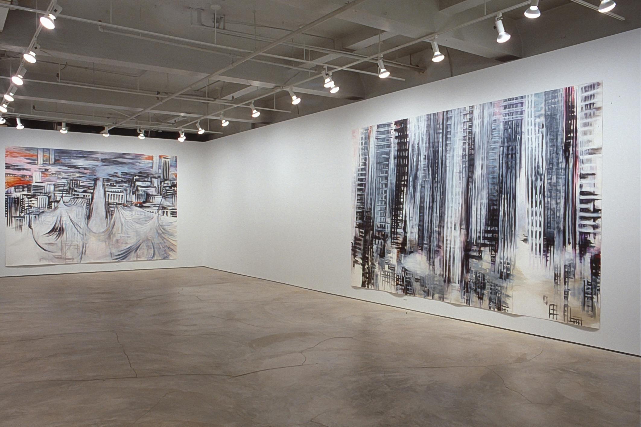 Two paintings are installed on the gallery walls. The one in the back shows an aerial view of a wide road running straight between buildings. The one in the front shows the bottoms of skyscrapers.  
