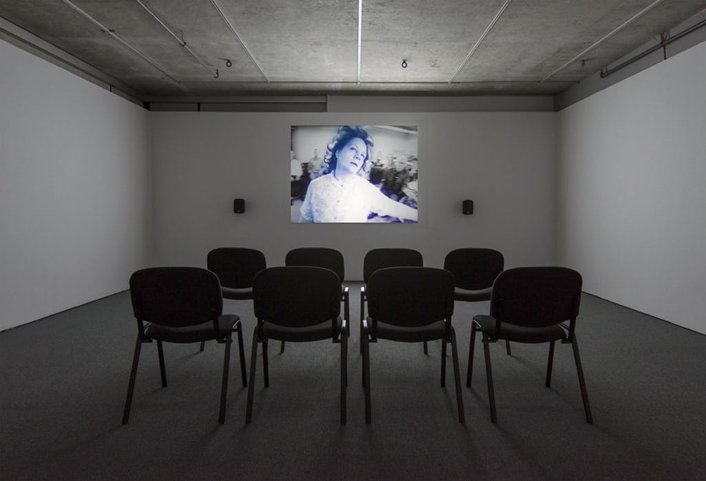 A single-channel video is projected on the wall. The video depicts a person in a white shirt dancing. Blue and purple lights frame her body. Black chairs are aligned on the floor toward the video. 