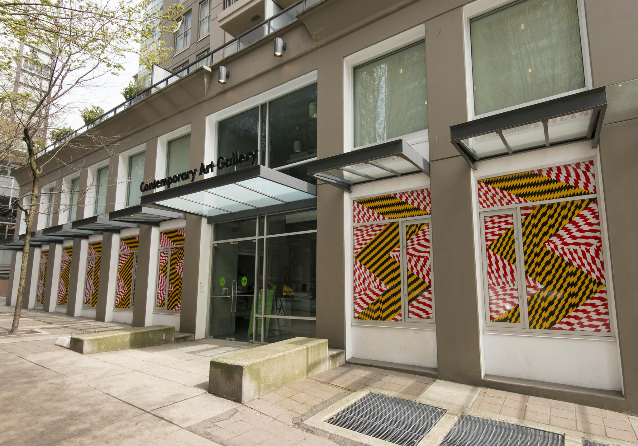 Exterior image of CAG with red-and-white and black-and-yellow caution tape installed across the ground floor façade windows. The tape forms zig-zag lines and shapes, covering each window.
