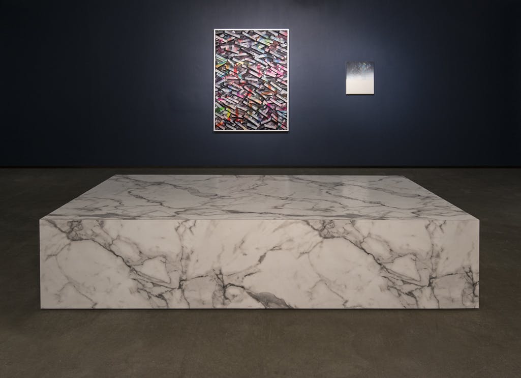 A large, rectangular sculpture resembling a marble plinth is installed in the middle of a gallery floor. Mounted on the dark blue wall behind are two, two-dimensional abstract artworks. 