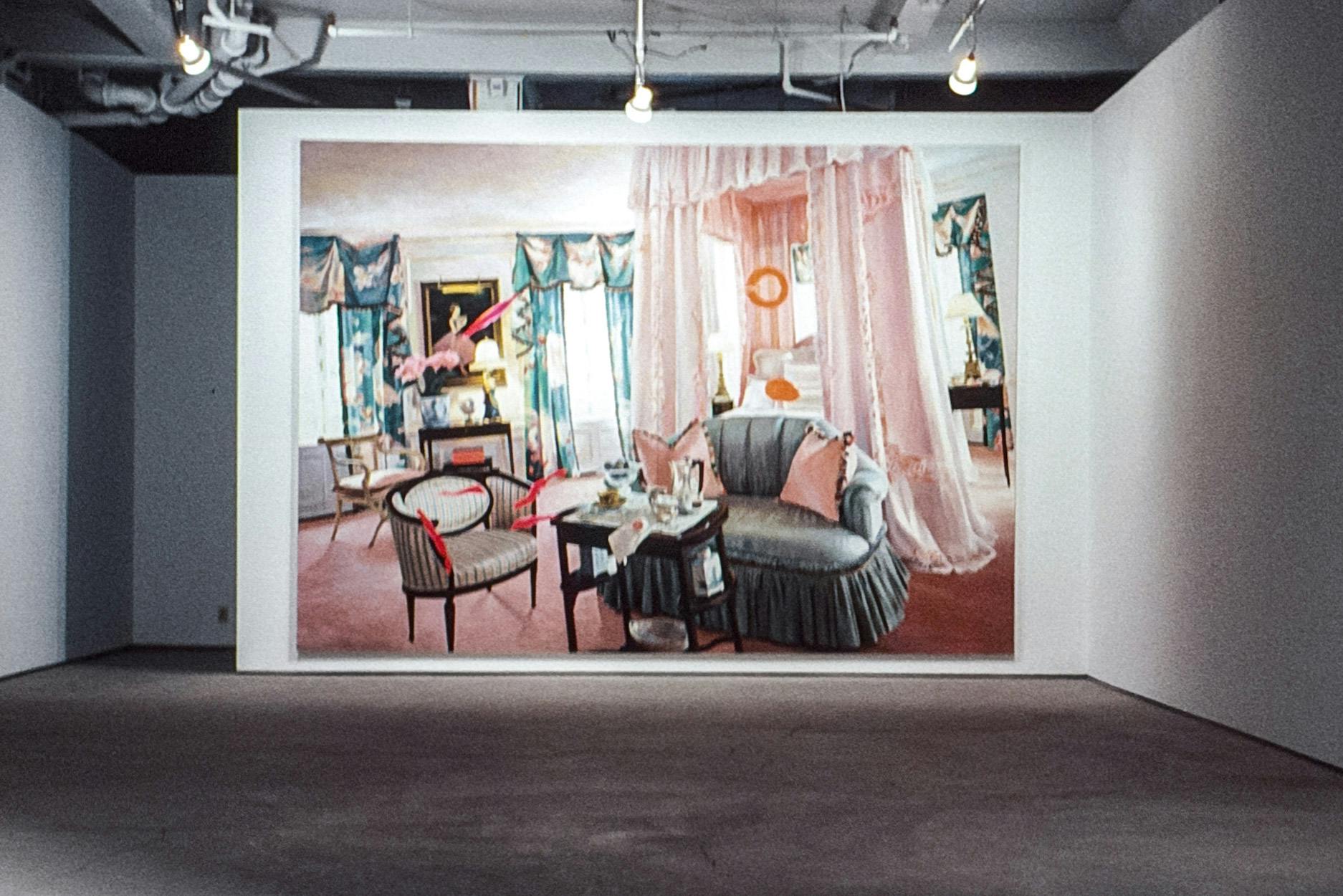 A painting in a gallery space nearly covers the wall it is mounted on. The painting shows a bright pink and blue bedroom with lush furnishings and is painted at a slightly distorted angle.