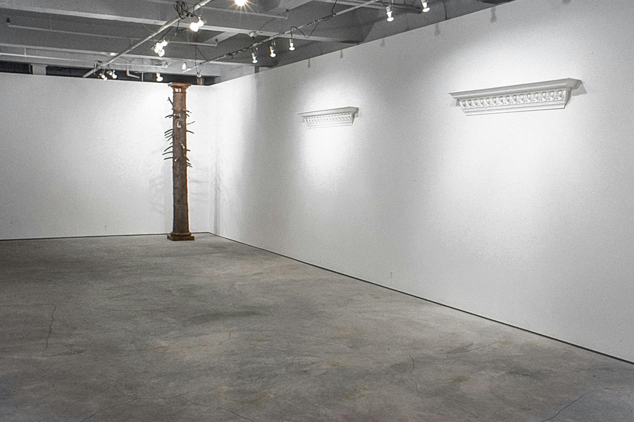 In a gallery, there are 2 ornate crown mouldings with partway up one wall, with small spaces in them where eggs rest. By another wall, there is a column made of a tree trunk with partial branches. 