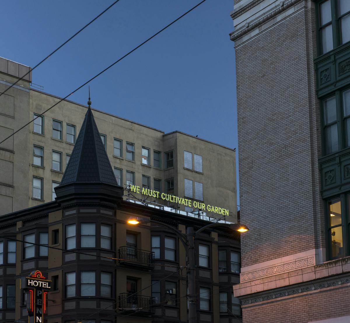 Nathan Coley’s work installed on the top loof of the Pennsylvania Hotel on East Hastings St. The Illuminated text reads “WE MUST CULTIVATE OUR GARDEN” in yellow-green light in the late evening sky.