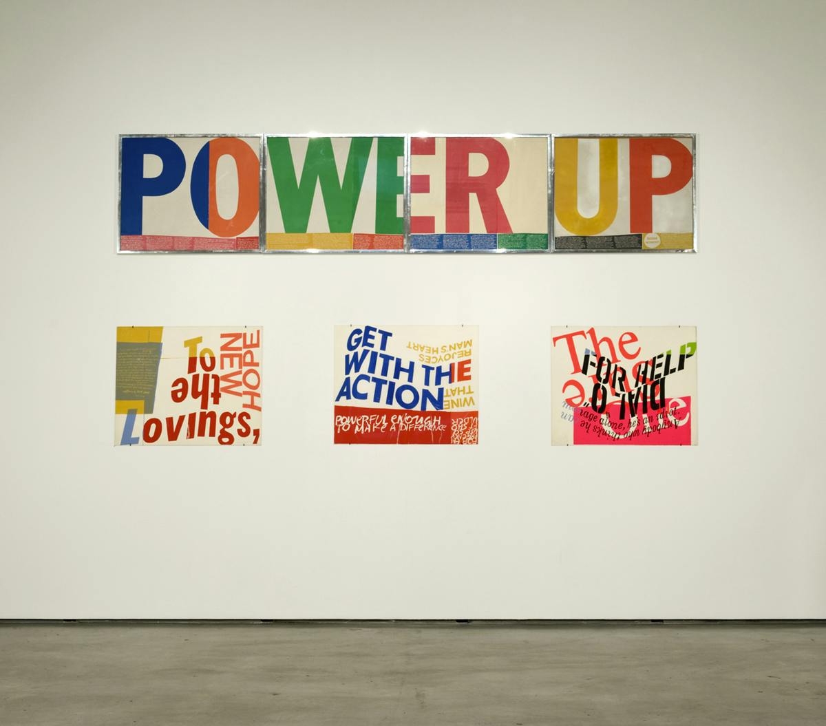 On a white gallery wall, a silver-framed colourful sign which reads “POWER UP” is mounted on the top. Below it, three other signs are mounted. One of them reads “GET WITH THE ACTION.”