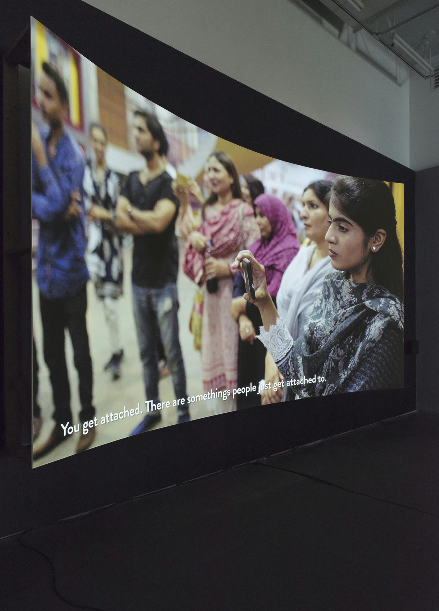 An angled view of a projector screen showing an image of a group of people standing, one is holding a phone. The subtitles read: “You get attached. There are somethings people just get attached to.”