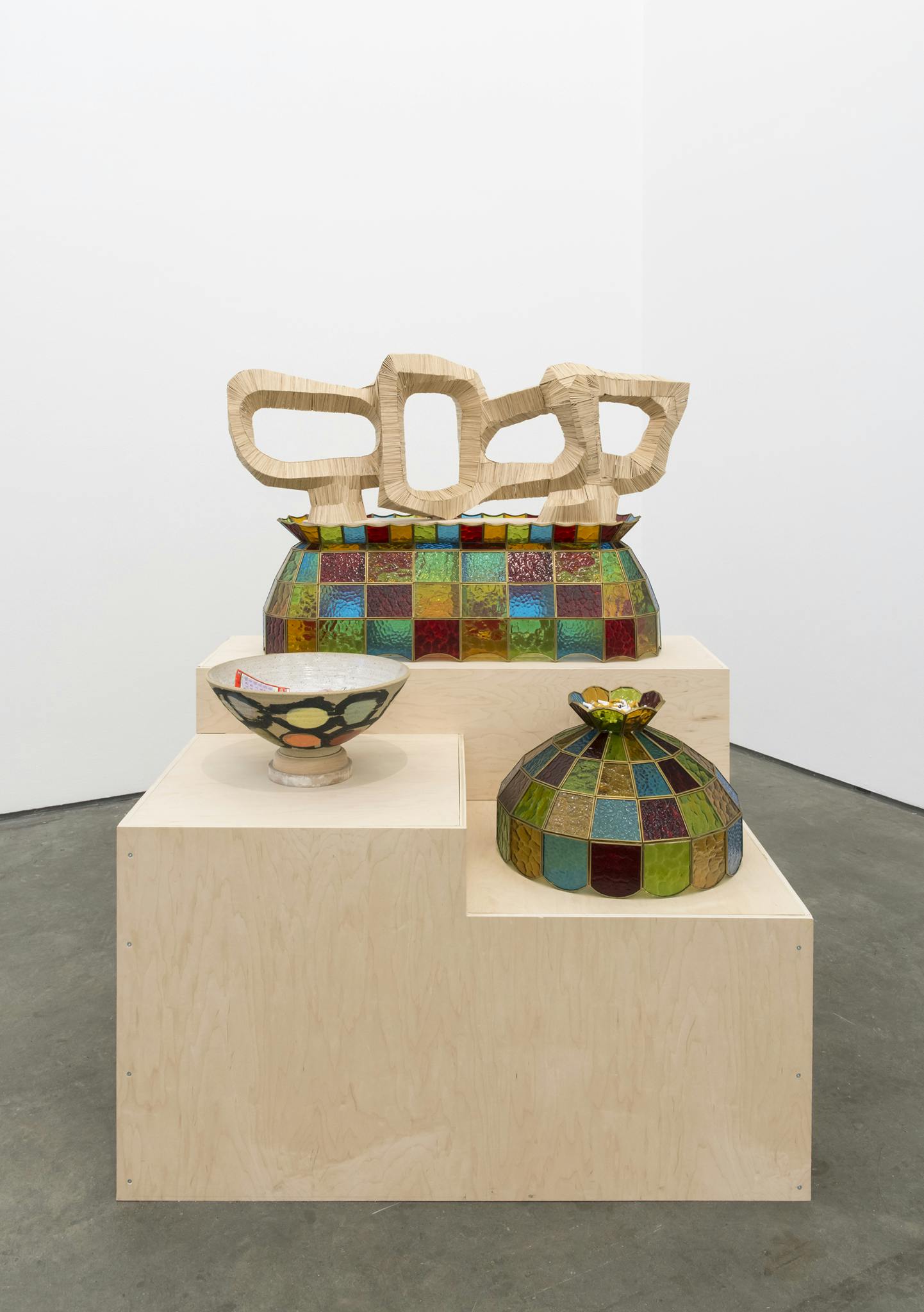 A ceramic bowl, wooden sculpture and two colourful glass lampshades sit on a three tier plinth made out of light wood.