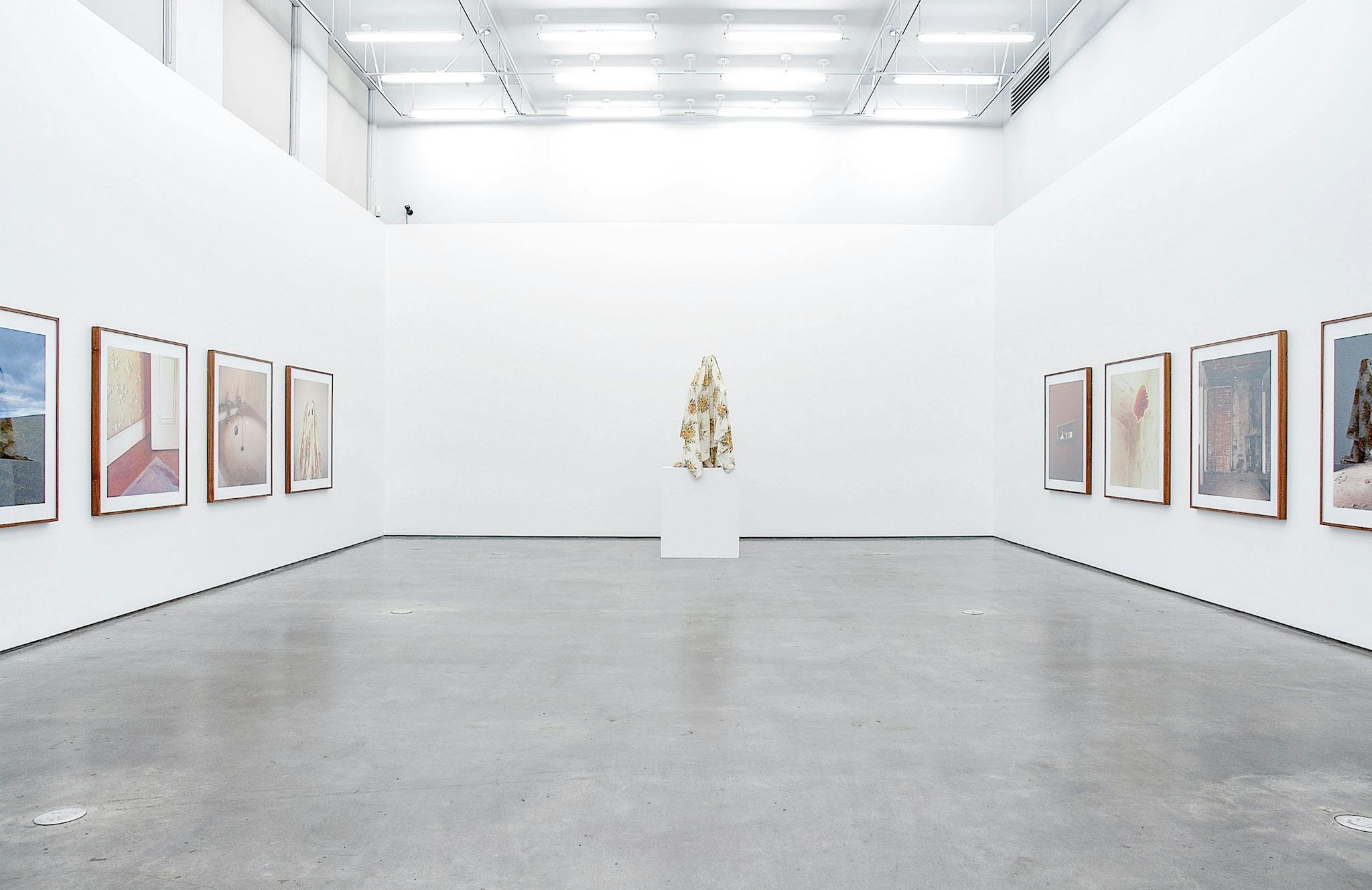 Three visible walls in a gallery. On two walls, there are large colour photos in wood frames. In front of the other wall is a large sculpture of a figure covered in a white sheet, resting on a plinth.
