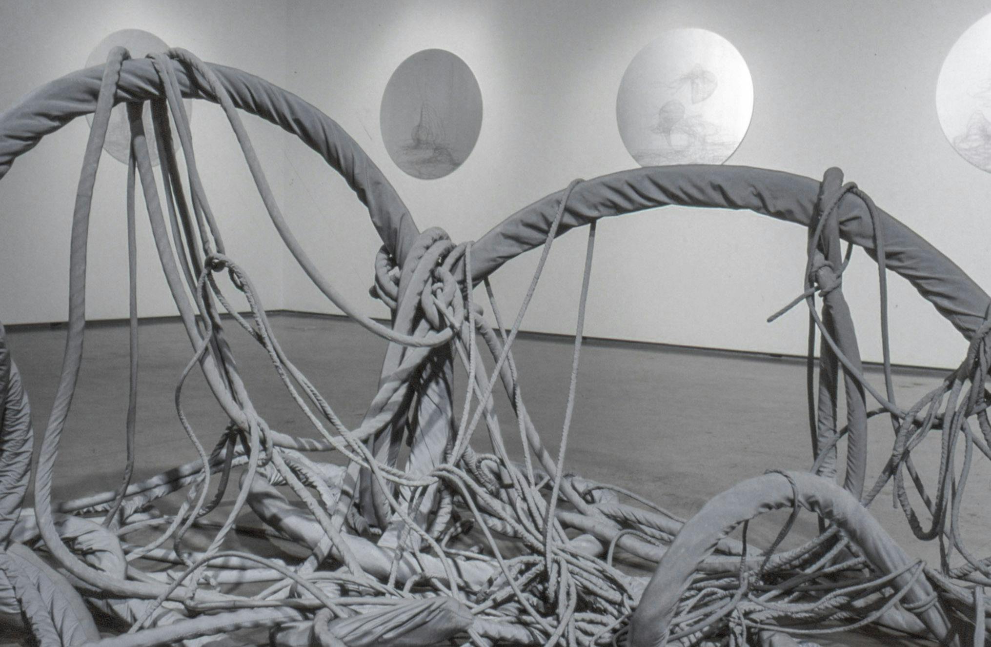 A large grey sculpture that looks like a tangled fishing net is placed in the middle of the gallery floor. Four circular shaped sculptures that resemble white marble are mounted on the walls. 