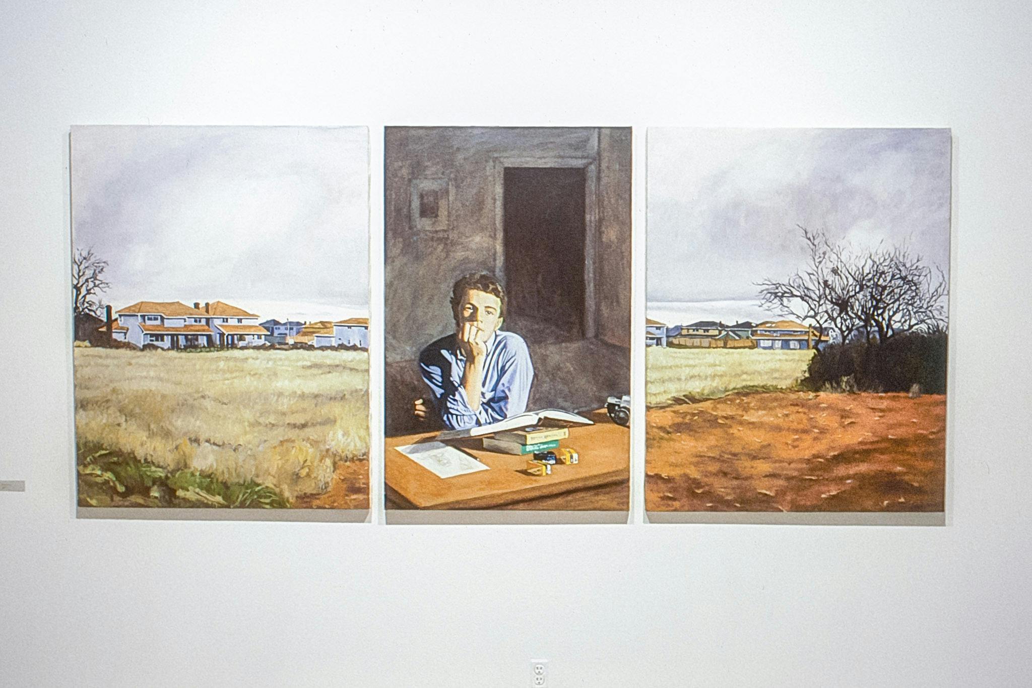 3 paintings mounted on a white wall. One shows a yellow field with houses in the background. One shows a person sitting at a desk with a book. One shows an orange field with houses in the distance. 