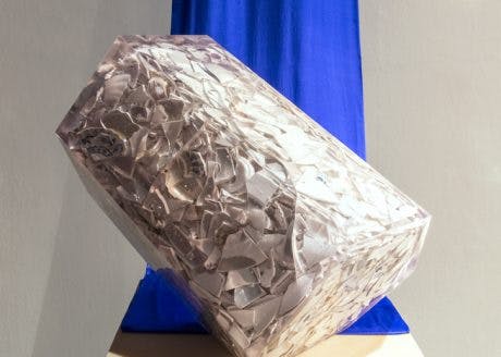 A clear, block-like sculpture of encased broken ceramic pieces installed on a plinth. A royal blue cloth hangs from the ceiling and drapes behind the sculpture. 