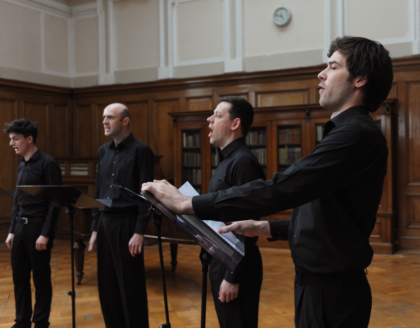 Four people in black outfit singing in a study. Each of them stands up straight with their mouth wide open. 